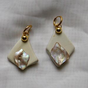 May include: A pair of earrings featuring off-white, diamond-shaped ceramic pieces adorned with iridescent, pearlescent shell fragments. Each earring is suspended from a gold-toned metal hoop with a small, polished gold-toned spherical bead.