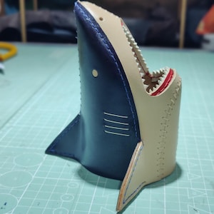 Puede incluir: Un portalápices artesanal de cuero con forma de tiburón. El portalápices es azul marino con una boca beige y detalles rojos. La boca del tiburón está abierta, revelando dientes. Las aletas son triangulares.