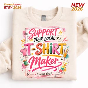 May include: Cream-colored sweatshirt with a colorful graphic that reads "Support Your Local T-Shirt Maker." The design includes a pink t-shirt, a spool of thread, and a color palette. The text "NEW 2026" is in the upper right corner.