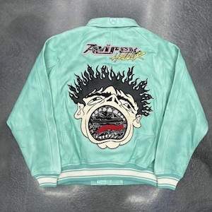 Könnte beinhalten: Hellgrüne Lederjacke mit großem Grafikdesign auf dem Rücken. Die Grafik zeigt ein schreiendes Gesicht mit schwarzen Haaren und roter Zunge. Der Text "Avirex Hellstar" befindet sich über der Grafik. Weiß-hellgrün gestreifte Bündchen und Saum.