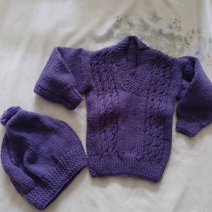 Puede incluir: Un conjunto de punto morado compuesto por un suéter y un gorro a juego. El suéter tiene un patrón de punto de cable y un cuello en V. El gorro tiene un pompón en la parte superior.