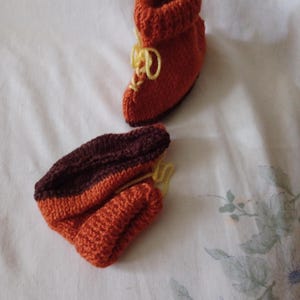 May include: A pair of knitted baby booties in orange and brown with yellow laces. The booties are folded and laid out on a white surface with a floral pattern.