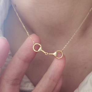 May include: A delicate gold-colored necklace featuring a horse bit pendant. The necklace is displayed against a soft, neutral background, highlighting the jewelry's intricate design and shine. The pendant is a classic equestrian-inspired design.