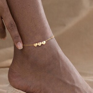May include: A gold-coloured anklet with four heart-shaped charms, each with a letter. The delicate anklet is worn on the ankle. The letters on the charms appear to spell a name or word. The anklet is a piece of jewellery.