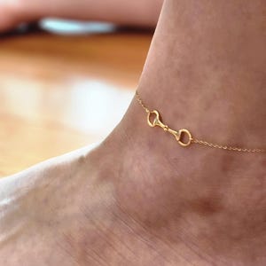 Op de afbeelding: Een delicate goudkleurige enkelband met een paardenbit-ontwerp. De enkelband is een fijne ketting met een kleine, gedetailleerde bit-bedel. De sieraden worden op de enkel gedragen.