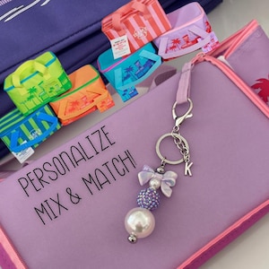 May include: A light purple pencil case with the text "PERSONALIZE MIX & MATCH!" and a pink zipper. A keychain with a pearl bead, purple rhinestone bead, and a silver initial "K" hangs from the case. Small colorful tote bags are scattered around the case.