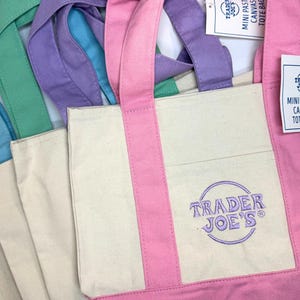 May include: Mini pastel canvas tote bags in shades of pink, purple, green, and blue. Each bag has a cream-colored body with a colored bottom and handles. The Trader Joe's logo is embroidered on the front.