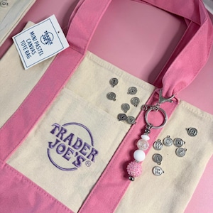 May include: A mini pastel canvas tote bag with pink handles and accents. The bag features the Trader Joe's logo in purple. A keychain with white and pink beads and charms hangs from the bag. Small silver charms are scattered around the bag.