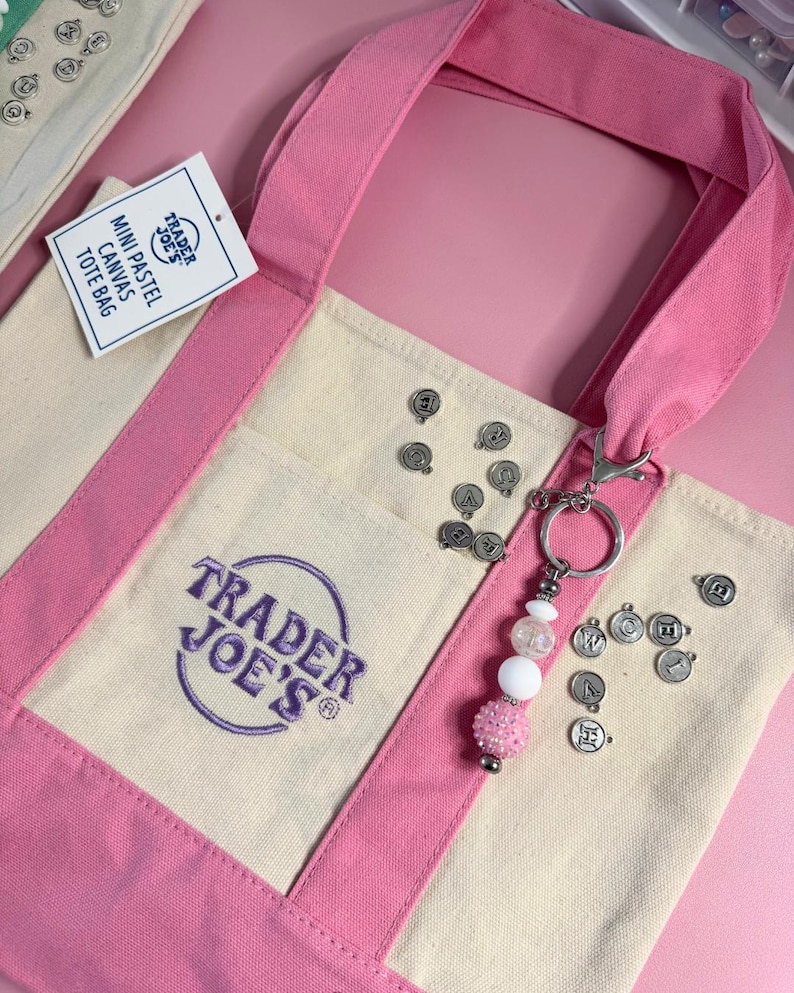 May include: A mini pastel canvas tote bag with pink handles and accents. The bag features the Trader Joe's logo in purple. A keychain with white and pink beads and charms hangs from the bag. Small silver charms are scattered around the bag.