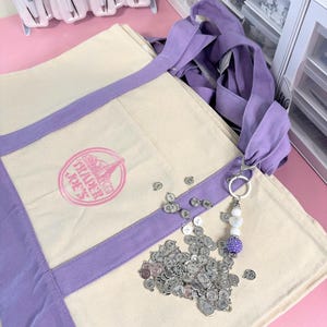 May include: A canvas tote bag with lavender accents and the Trader Joe's logo. A keychain with a purple beaded charm and silver letter charms is next to the bag. Pens with decorative tops are visible in the background.