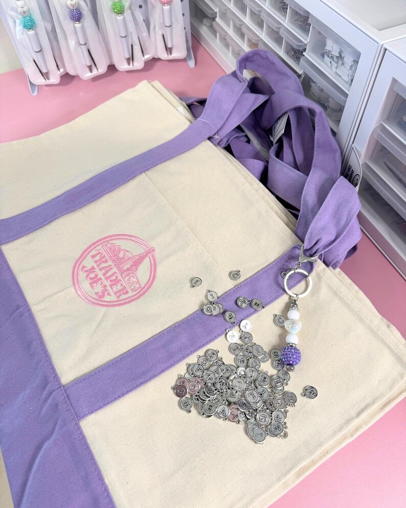 May include: A canvas tote bag with lavender accents and the Trader Joe's logo. A keychain with a purple beaded charm and silver letter charms is next to the bag. Pens with decorative tops are visible in the background.
