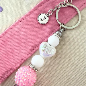 May include: A silver keychain with a round charm featuring the letter "J". The keychain has a beaded design with a pink, sparkly ball, white beads, and a heart-shaped bead with a bow design. The background is a pink strap.