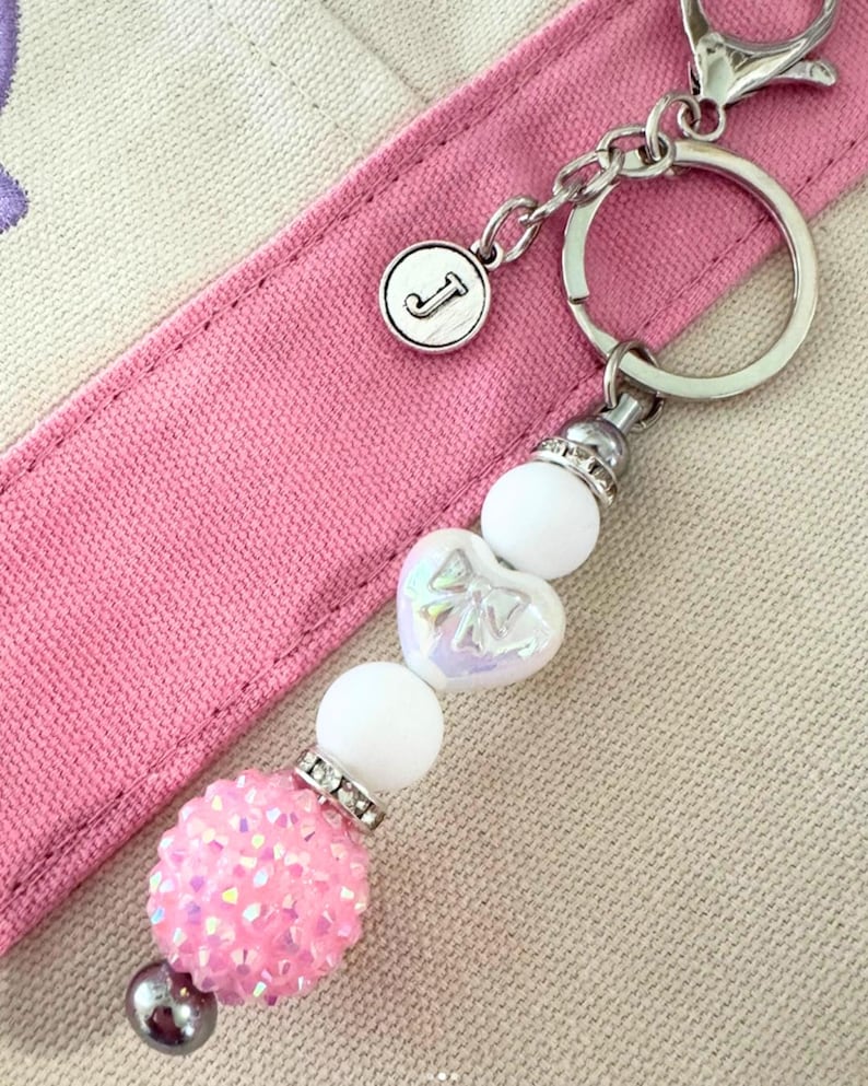 May include: A silver keychain with a round charm featuring the letter "J". The keychain has a beaded design with a pink, sparkly ball, white beads, and a heart-shaped bead with a bow design. The background is a pink strap.