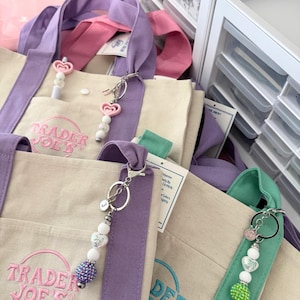 May include: Beige canvas tote bags with purple, pink, and green straps. The bags have the "Trader Joe's" logo in pink and teal. Keychains with heart-shaped beads and a small initial charm are attached to the bags.
