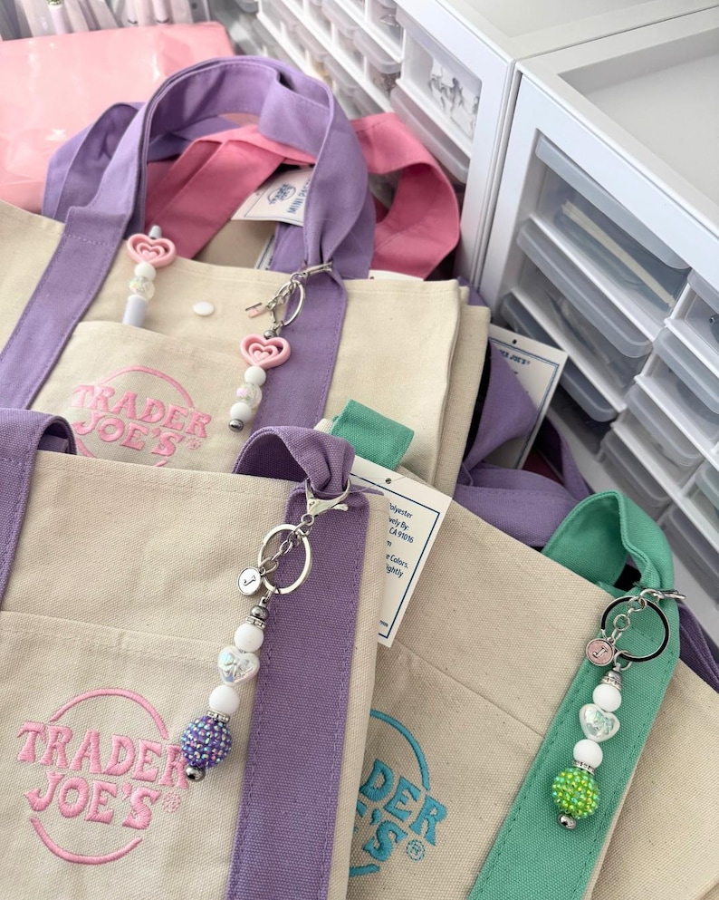 May include: Beige canvas tote bags with purple, pink, and green straps. The bags have the "Trader Joe's" logo in pink and teal. Keychains with heart-shaped beads and a small initial charm are attached to the bags.