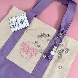 May include: A canvas tote bag with lavender accents and handles. The bag features the Trader Joe's logo in pink and a tag that reads "Mini Pastel Canvas Tote Bag." A beaded keychain with silver charms hangs from the handle.