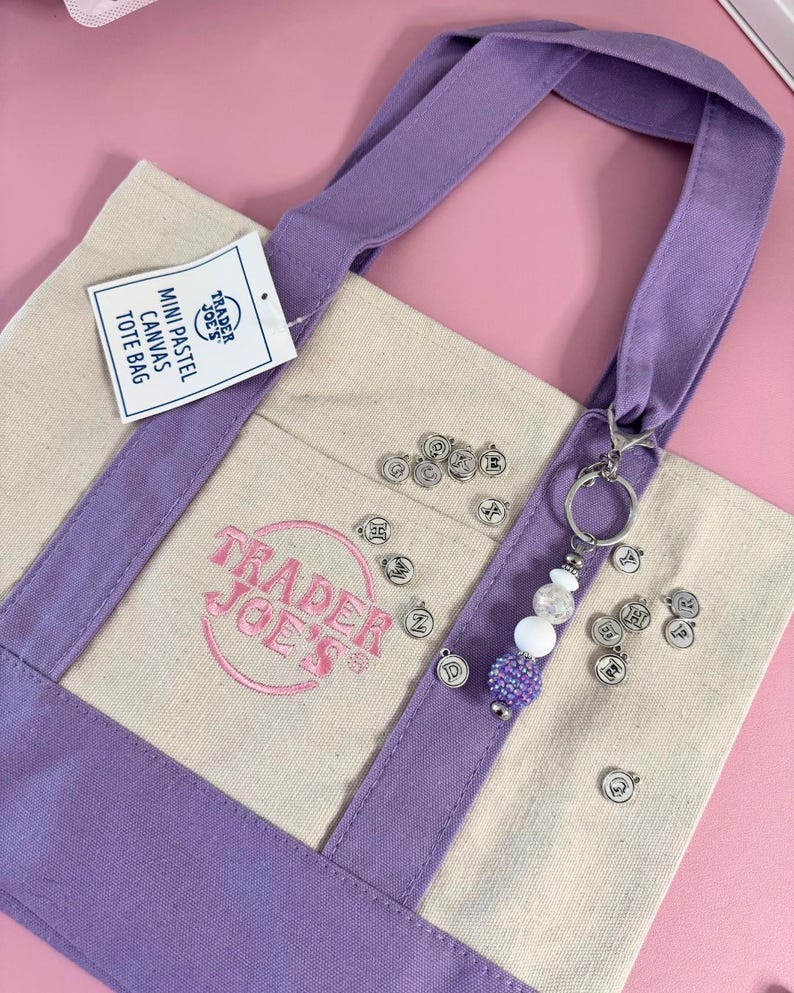 May include: A canvas tote bag with lavender accents and handles. The bag features the Trader Joe's logo in pink and a tag that reads "Mini Pastel Canvas Tote Bag." A beaded keychain with silver charms hangs from the handle.