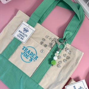 May include: A mini pastel canvas tote bag with green accents and handles. The bag features the Trader Joe's logo and a tag that reads "MINI PASTEL CANVAS TOTE BAG." A keychain with letter charms and beads hangs from the handle.