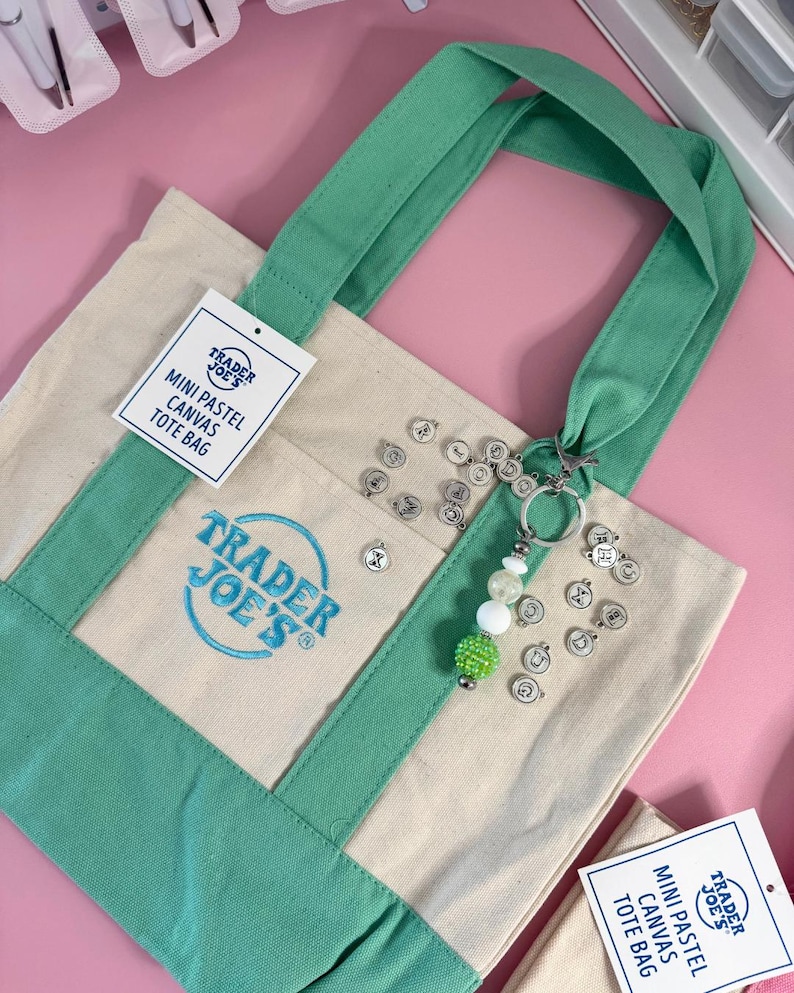 May include: A mini pastel canvas tote bag with green accents and handles. The bag features the Trader Joe's logo and a tag that reads "MINI PASTEL CANVAS TOTE BAG." A keychain with letter charms and beads hangs from the handle.