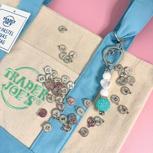 May include: A light blue and beige canvas tote bag with the Trader Joe's logo in green. A silver keychain with white and turquoise beads hangs from the handle. Small silver charms with letters are scattered around the bag.