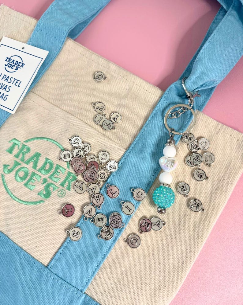 May include: A light blue and beige canvas tote bag with the Trader Joe's logo in green. A silver keychain with white and turquoise beads hangs from the handle. Small silver charms with letters are scattered around the bag.