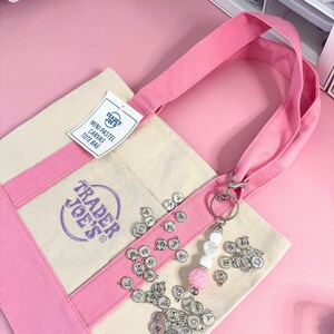 May include: A canvas tote bag with pink accents and handles, featuring the Trader Joe's logo. The bag has a tag that reads "MINI PASTEL CANVAS TOTE BAG." Silver letter charms and a beaded keychain with a pink pom-pom are attached.
