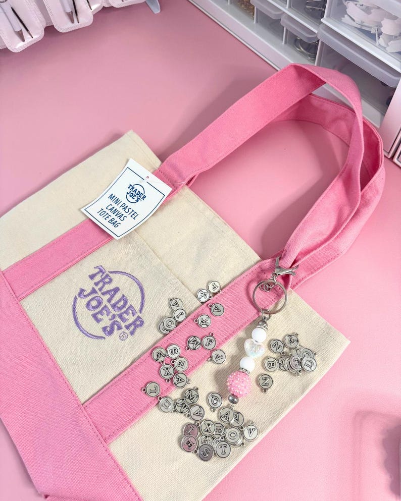 May include: A canvas tote bag with pink accents and handles, featuring the Trader Joe's logo. The bag has a tag that reads "MINI PASTEL CANVAS TOTE BAG." Silver letter charms and a beaded keychain with a pink pom-pom are attached.