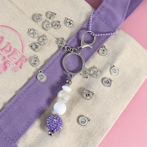 May include: A keychain with a silver ring and lobster clasp, featuring a beaded charm with white and purple beads. Scattered around are small silver charms with letters. The keychain is on a canvas tote bag with a purple strap.