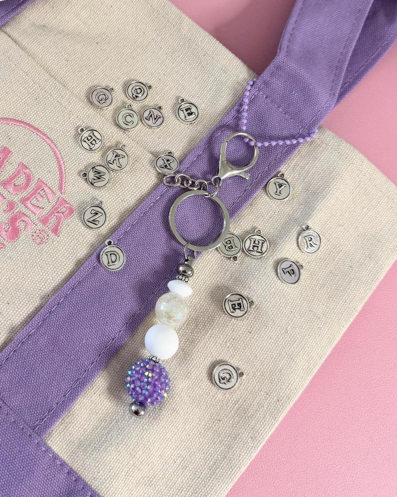 May include: A keychain with a silver ring and lobster clasp, featuring a beaded charm with white and purple beads. Scattered around are small silver charms with letters. The keychain is on a canvas tote bag with a purple strap.