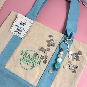 May include: A mini pastel canvas tote bag with light blue handles and accents. The bag features the Trader Joe's logo in green and a tag that reads "Mini Pastel Canvas Tote Bag." A keychain with beads and letter charms is attached.