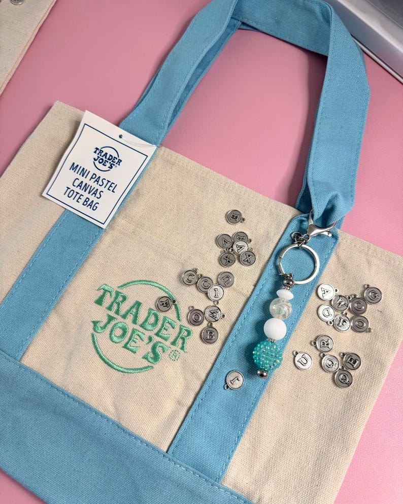 May include: A mini pastel canvas tote bag with light blue handles and accents. The bag features the Trader Joe's logo in green and a tag that reads "Mini Pastel Canvas Tote Bag." A keychain with beads and letter charms is attached.