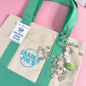 May include: A mini pastel canvas tote bag with green handles and trim. The bag features the Trader Joe's logo in blue. A keychain with letter charms and decorative beads hangs from the handle. The background is a light pink.
