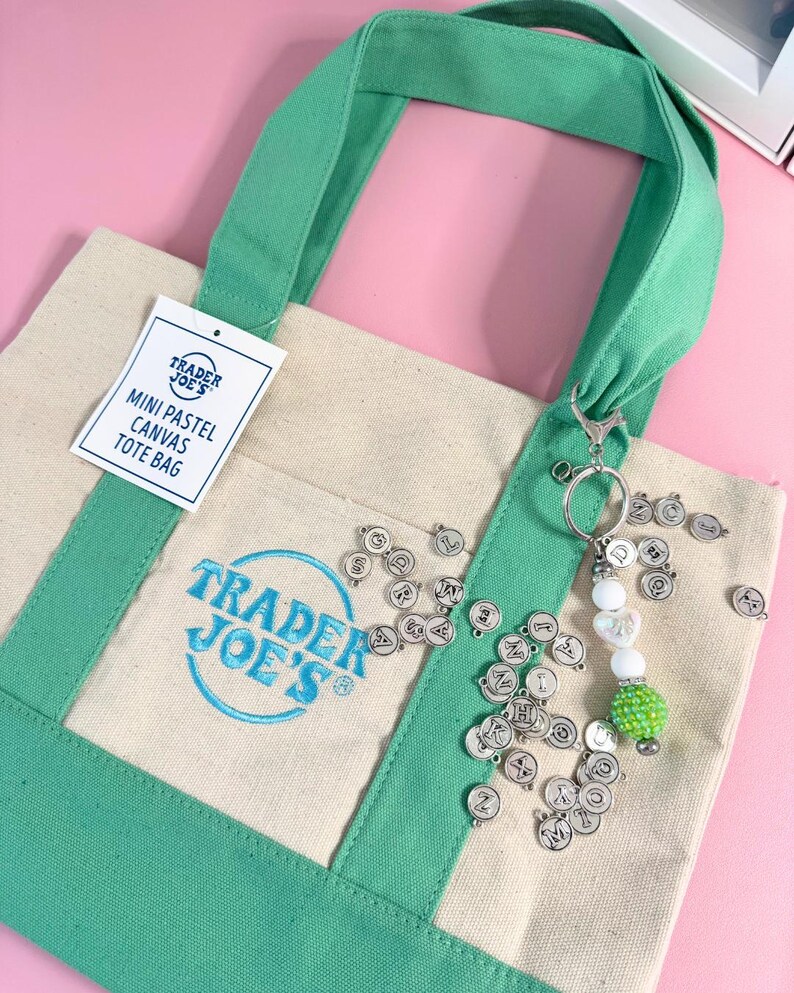 May include: A mini pastel canvas tote bag with green handles and trim. The bag features the Trader Joe's logo in blue. A keychain with letter charms and decorative beads hangs from the handle. The background is a light pink.