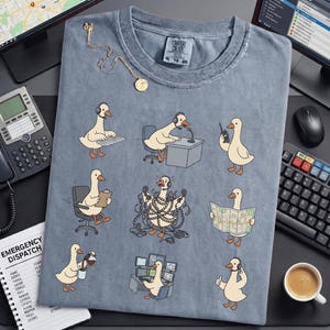 May include: A blue-grey t-shirt featuring cartoon geese in various emergency dispatch roles. The geese are depicted using a computer, radio, phone, and map. The shirt has the text "COMFORT COLORS" and size information.