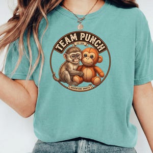 May include: A teal-colored t-shirt featuring a circular graphic with two cartoon monkeys. The graphic includes the text "TEAM PUNCH EST. 2020" and "ADVENTURE AWAITS" in a brown and tan color scheme. The shirt has short sleeves.
