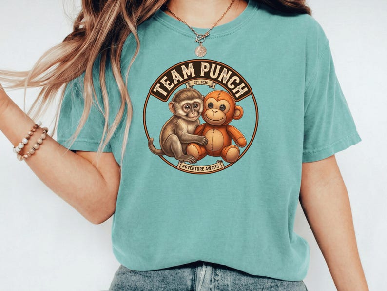 May include: A teal-colored t-shirt featuring a circular graphic with two cartoon monkeys. The graphic includes the text "TEAM PUNCH EST. 2020" and "ADVENTURE AWAITS" in a brown and tan color scheme. The shirt has short sleeves.
