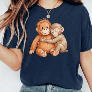 May include: Navy blue t-shirt featuring a watercolor illustration of two hugging monkeys. One monkey is orange, the other is tan. The design is centered on the shirt, creating a heartwarming image. The shirt is made of a soft material.