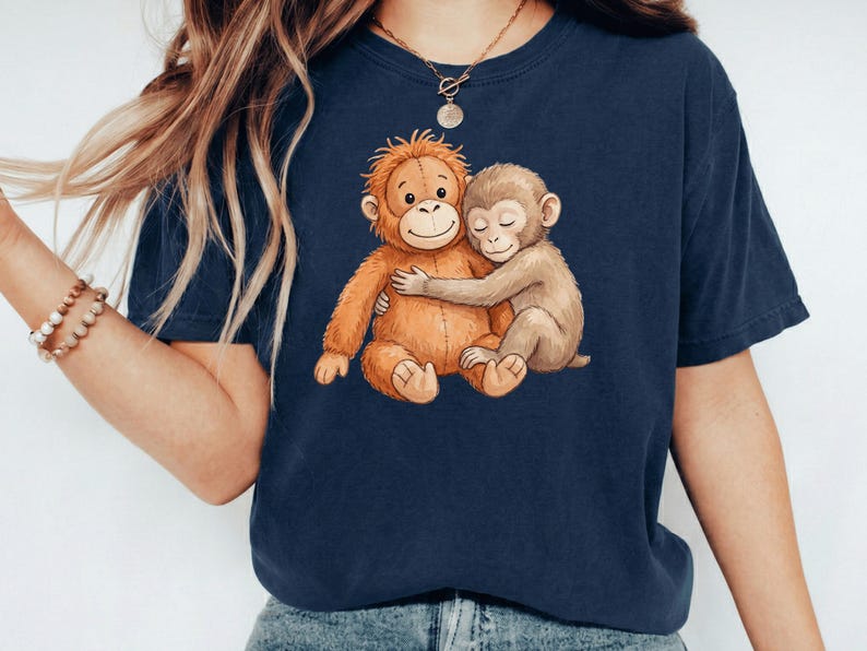 May include: Navy blue t-shirt featuring a watercolor illustration of two hugging monkeys. One monkey is orange, the other is tan. The design is centered on the shirt, creating a heartwarming image. The shirt is made of a soft material.
