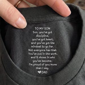 Op de afbeelding: Donkergrijs t-shirt met witte tekst. De tekst luidt "TO MY SON" gevolgd door een oprechte boodschap van "DAD". Het shirt wordt vastgehouden door een hand met een gouden ring.