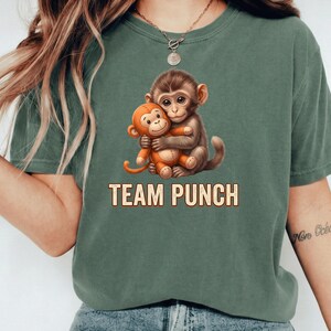 May include: Green t-shirt featuring a graphic of two cartoon monkeys hugging. The text "TEAM PUNCH" is printed below the monkeys in a tan color. The shirt is a solid green color.
