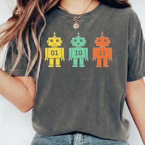 May include: Dark grey t-shirt featuring three colorful robot graphics. Each robot is a different color: yellow, green, and orange, with the numbers 01, 10, and 11 displayed on their chests. The robots have antenna and square feet.