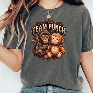 May include: Gray t-shirt featuring a graphic of two cartoon monkeys hugging, with the text "TEAM PUNCH" in a decorative emblem. The monkeys are brown and orange, and the emblem is brown and gold.