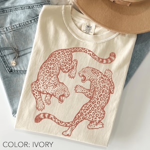 Puede incluir: Camiseta de color marfil con una ilustración marrón rojizo de dos leopardos. Uno salta, el otro ruge. La camiseta es de la marca Comfort Colors. El texto 'COLOR: IVORY' está en la parte inferior de la imagen.