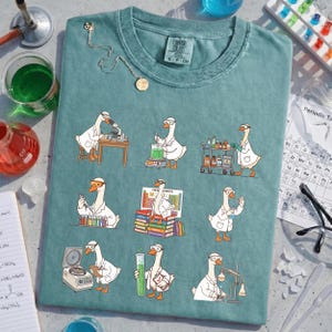 Peut inclure: T-shirt bleu sarcelle avec des oies de dessin animé en blouses de laboratoire effectuant des expériences scientifiques. Les oies utilisent des microscopes, des béchers et d'autres équipements de laboratoire. Le t-shirt porte une petite étiquette indiquant "COMFORT COLORS S-P CH".
