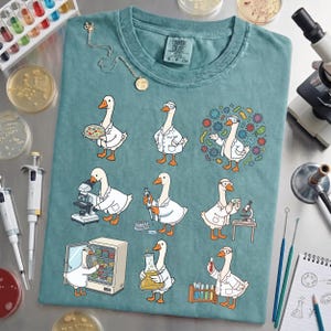 Könnte beinhalten: Türkisfarbenes T-Shirt mit Cartoon-Gänsen in Laborkitteln, die wissenschaftliche Aktivitäten ausüben. Die Gänse werden mit Mikroskopen, Bechern und Petrischalen dargestellt. Das Shirt hat einen Rundhalsausschnitt.