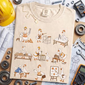 May include: A beige t-shirt featuring a graphic print of nine cartoon geese dressed as construction workers and engineers, performing various tasks like drafting, measuring, and building. The shirt has a crew neck and a label reading Canvas Jours XL.