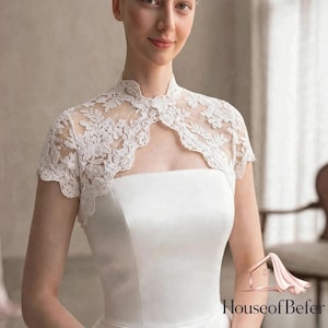May include: A white lace bolero jacket featuring short sleeves, a high neckline, and intricate floral embroidery. The garment is worn over a strapless white dress. The logo House of Befer is visible in the bottom right corner of the image.