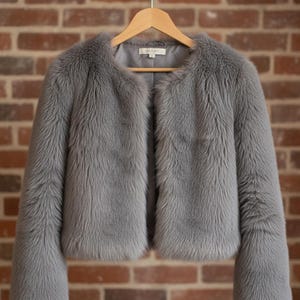 May include: A gray faux fur jacket with a round collar and long sleeves hangs on a wooden hanger. The jacket has a soft, textured appearance and a light gray lining. The background is a brick wall.