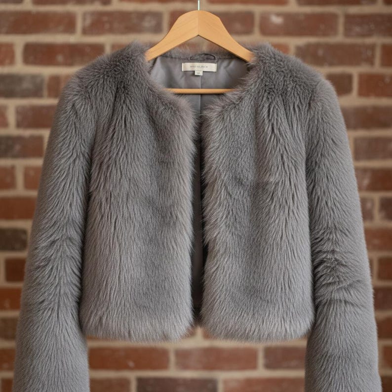 May include: A gray faux fur jacket with a round collar and long sleeves hangs on a wooden hanger. The jacket has a soft, textured appearance and a light gray lining. The background is a brick wall.