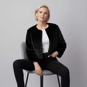 May include: A black faux fur jacket with a round neckline. The cropped jacket is worn over a white top and black jeans. The model is seated on a gray chair against a gray background.